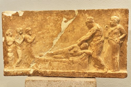 Votive relief of Asclepius The god bends down over the reclining sick woman, placing his hands on he