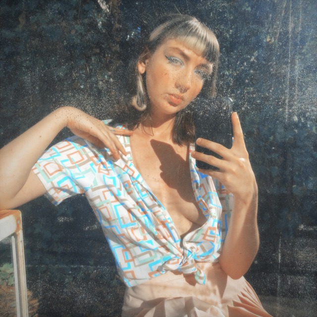 Peaches, a pale thin femme, posing in a mirror with her left hand resting on her collar bone, wearing a white button up shirt with pastel shapes on it un buttoned to reveal cleavage