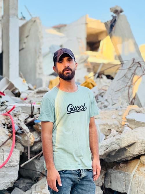 A photo of Hossam. He is a young man with a short cropped beard. He is wearing a navy blue New York Yankees baseball cap and a mint green t-shirt with the word 'Gucci' printed in black. He is standing amid the rubble of a building.