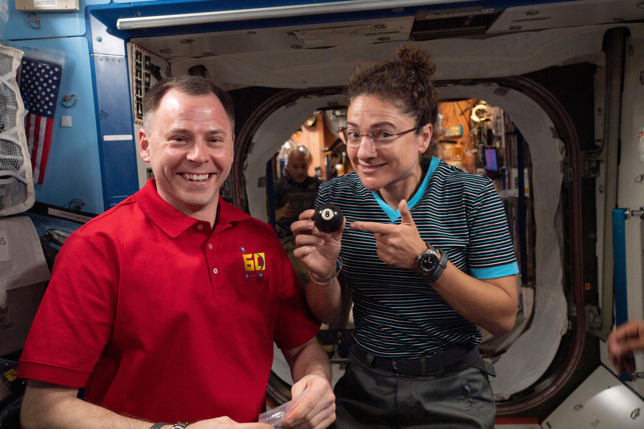 NASA astronauts Nick Hague and Jessica Meir hold up an 8-ball plushie and smile at the camera aboard the International Space Station, symbolizing their membership in the Astronaut Class of 2013. Credit: NASA