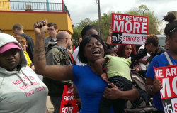 tranqualizer:  [photo: centered in the photo is a Black woman carrying a child in one arm with her other arm and fist raised. she is in the middle of chanting. there are others around her. a protest sign from the fast food strike in the background reads,