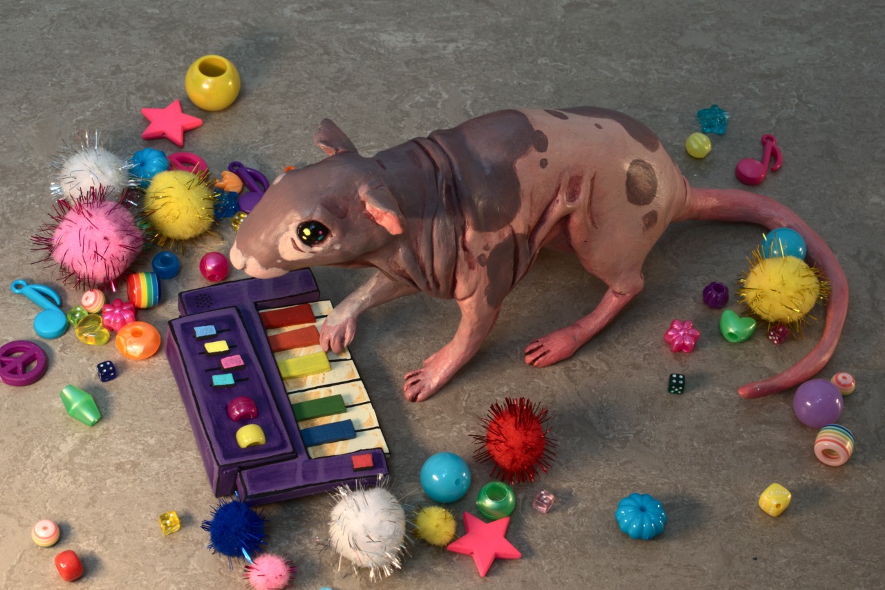 A photo of a hand-sized painted clay figurine of Neil the hairless rat, playing a small purple synthesizer. The photo is taken slightly from above, showing Neil's left side.  The knobs, wheels and black keys of the synth are in the colors of the rainbow. The figurine has its right forepaw raised, resting on the keys.  The figurine sits on a grey vinyl floor, and is surrounded by colorful glittery pompoms and plastic beads in a variety of shapes laid out around it. Some of the beads are shaped like stars and musical notes.