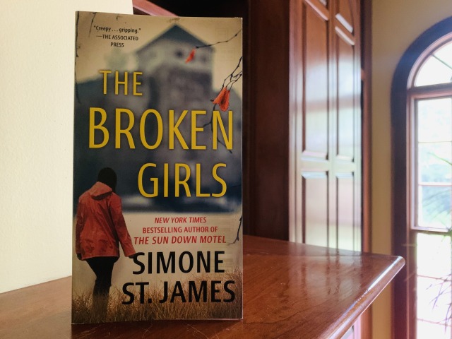 Shown is THE BROKEN GIRLS (with cover art depicting a young woman in a coat, beside a bare tree, approaching an out-of-focus building) on a wood mantel beside a window. Photo by AHS.