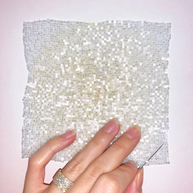 A painting with a yellow glowing circle in the centre, a white halo, then a pink background. It has an irregularly shaped square of white beads covering most of it. It has a sparkly ethereal look. 