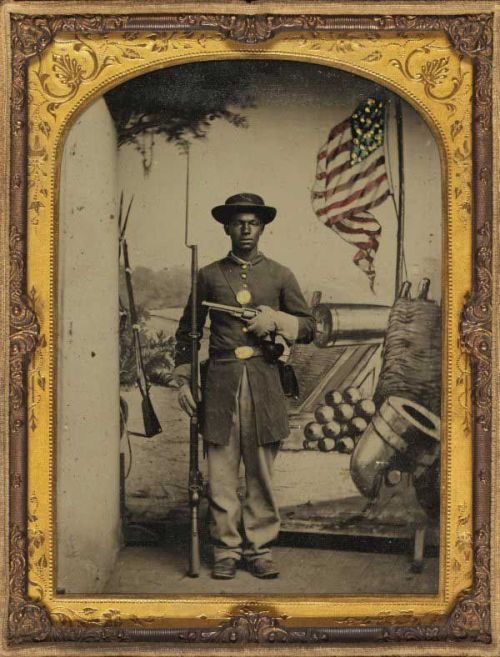 blackhistoryalbum:  African American soldier in Union uniform with a rifle and revolver posing in front of painted backdrop with American flag and artillery pieces. 
