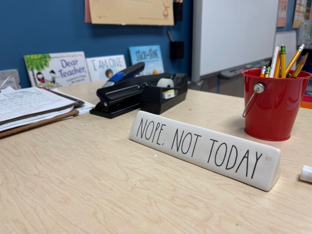 A dirty teacher's desk - notably minus any evidence of computer use, whatsoever. The subject in focus is a name card that says, simply, "NOPE, NOT TODAY" in all caps.