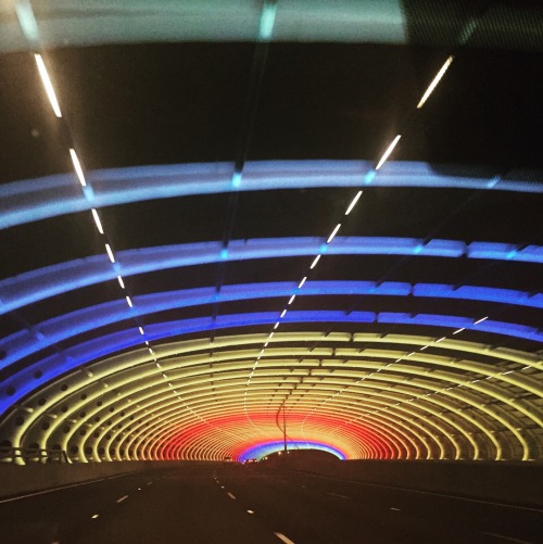 [ID: A photo of a tunnel at night. The inside is lit with arches glowing in blue, then yellow, then 