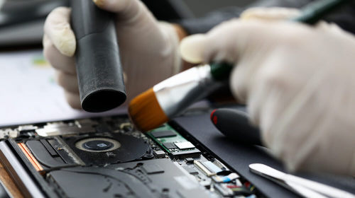 a person with gloved hands carefully brushing dust out of an open laptop and into a vacuum hose.