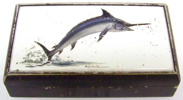 A cigarette box with a painted cover image on porcelain tile of a swordfish jumping out of the water. The artist's signature is below the image: Reinbold. The box is covered with gold-trimmed blue leather. 