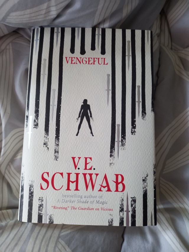 the cover of a hardback edition of vicious which is white with black stripes and daggers pointing down with a small silhouette of a woman in black in the centre. the title and authors name is in red. the book rests on grey and white patterned fabric