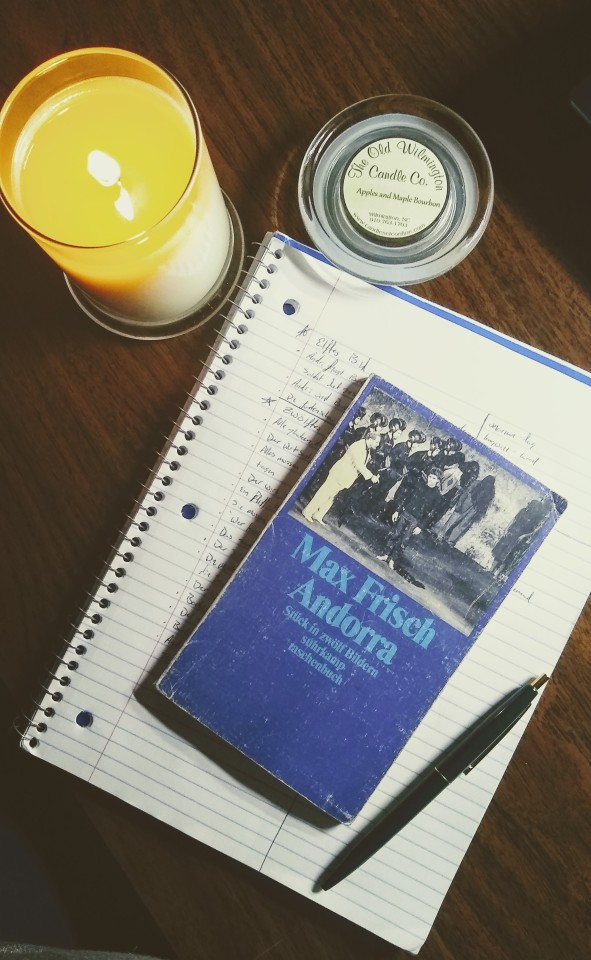 a photo of a pen and the book "andorra" by max frisch laying on top of a notebook. the book and notes are in german. there is a lit candle and its lid, showing that the scent is apple maple bourbon