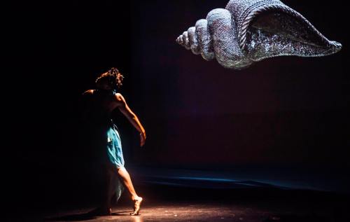 Image: A lone dancer is spotlighted on a stage with an image of a shell projected on a screen behind them.