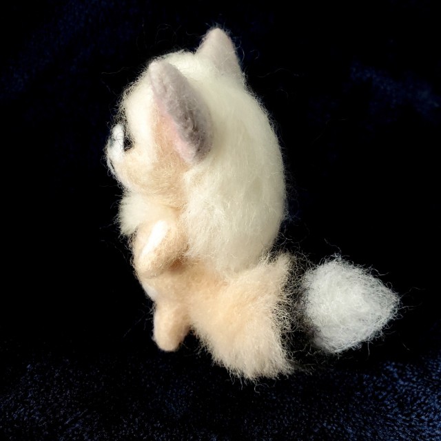 A photo of a needle felted sculpture of a fox-like creature named Menoes. She is peach colored with long, silver hair and white markings that look like bones. She is against a dark blue background.