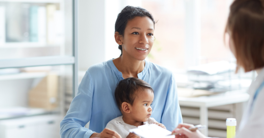 Image shows a young Latina woman with a baby, sitting across from a doctor, and smiling. 