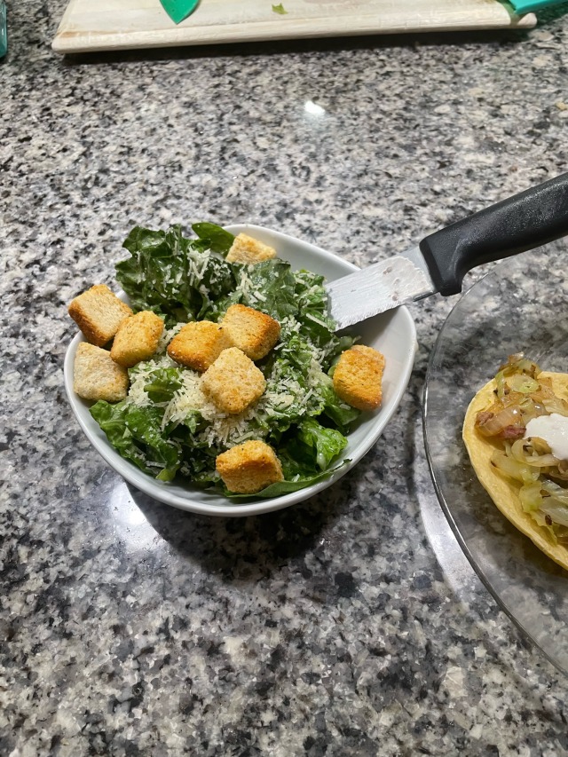 a white bowl on a granite countertop, it contains a Caesar salad with a large stake knife in it.