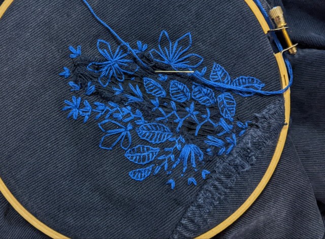 A blue pain of jeans with a rip in them, embroidery over with flowers and leaves in lighter blue.