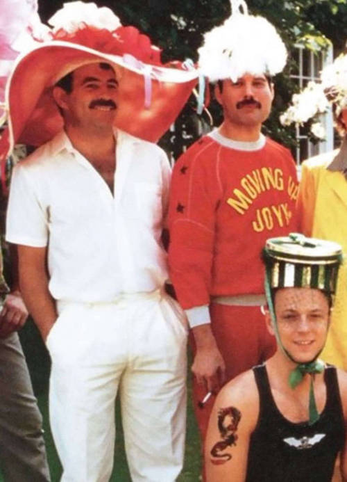 Cropped image of Freddie and Jim, two dark-haired mustachioed men, standing side-by-side in a group photo at a fancy-dress party. Jim is wearing a large, wide-brimmed pink hat, a white shirt and white pants. Freddie is wearing a white feathered hat, and a red tracksuit with the words 'MOVING UP JOYY' printed on it.
