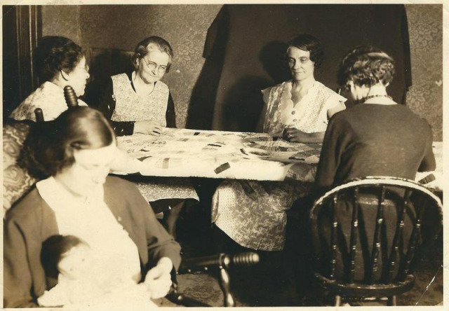 Black and white photograph of 5 women sitting around a table working on a quilt, one of the women in the foreground holding a doll.