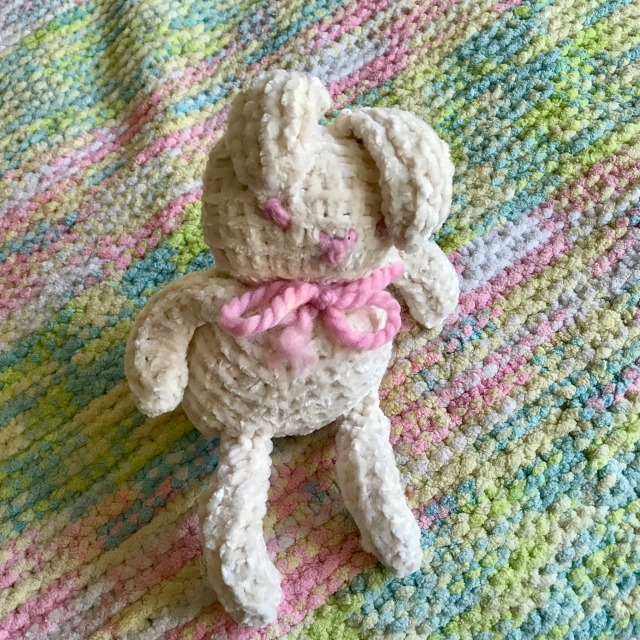 A knitted white bunny with pink eyes and a pink bow rests upon a pastel multicolored knitted blanket