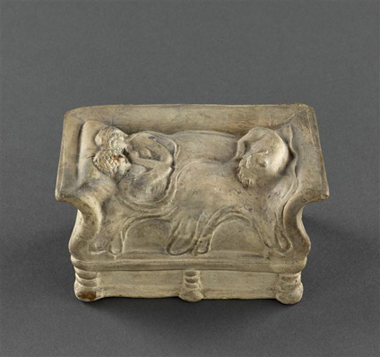 A terracotta model of two people embracing in bed, nude but under a blanket. One holds the other's chin. At the foot of the bed, a dog is curled up asleep. The same object as the last photo from a different angle.