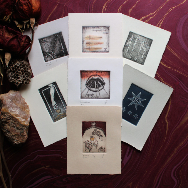 A photo of seven 4x5” hand-embellished etchings fanned out on a wine colored sheet of marbled paper. The imagery of the etchings resembles small, enigmatic tarot cards.