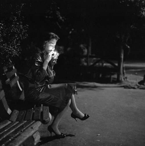 Hulton Archive. A young woman lighting a cigarette as she sits on a park bench at night. New York City. 1957. Nudes &amp; Noises  