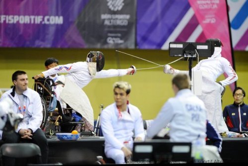 [ID: a wheelchair epee fencer lunging at his opponent, while other wheelchair fencers or coaches sit