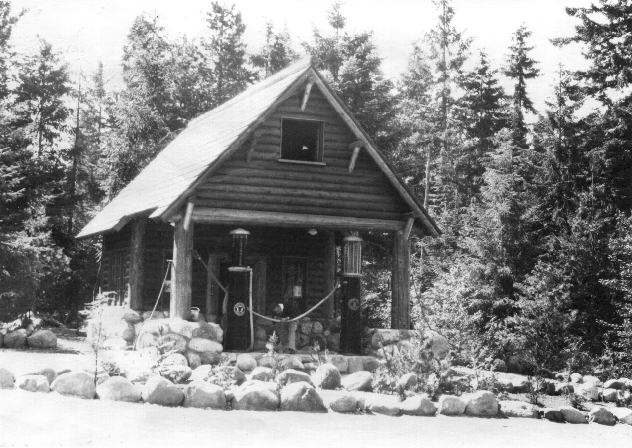 Black and white historic photo of a small log building with a stone foundation and gable roof. The roof extends out over two tall gas pumps on a stone island.  A planting bed ringed with large stones is in front of the building.