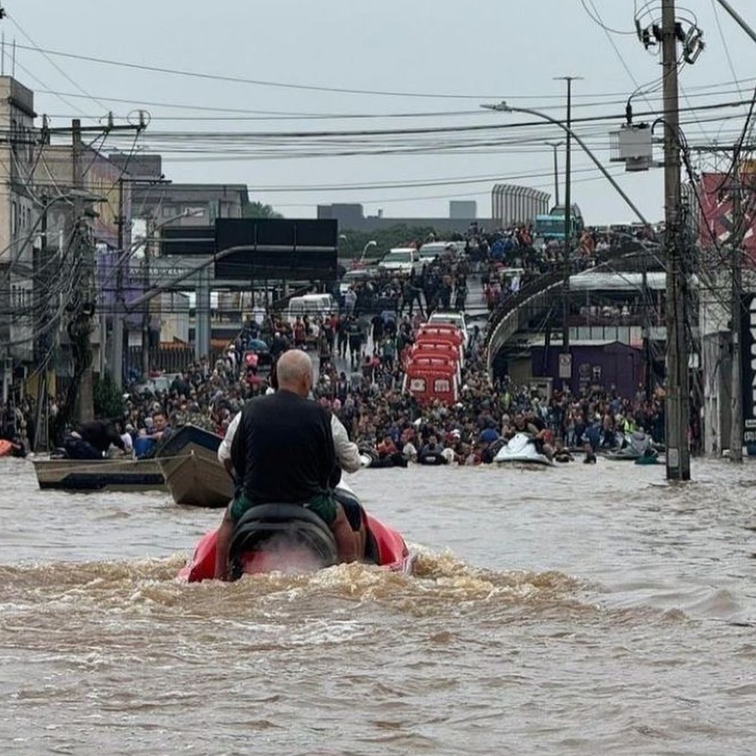 Picture taken in Canoas. A man pilots a jet ski towards an above water viaduct, where dozens of rescued people are awaiting. Another jet ski and two boats are visible. On the viaduct, four ambulances and many police cars are stationed.