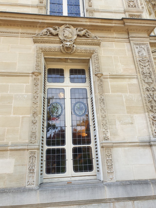 an outside view of a window euh the posts carved with flowers. above it is the face of a figure with a laurel crown, the name has faded to illegibility