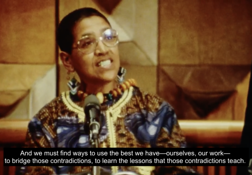 Audre Lorde stands at a podium accepting her award and appointment as State Poet of New York for 1991-1993. She is dressed formally in a colorful tunic. She continues, "And we must find ways to use the best we have--ourselves, our work--to bridge those contradictions, to learn the lessons that those contradictions teach."