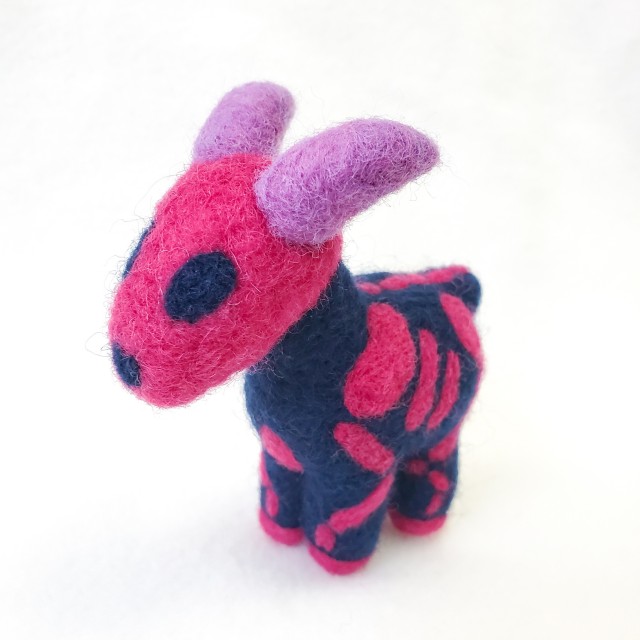 A photo of a dark blue needle felted ewe with purple horns and a pink skeleton pattern. The colors are in reference to the bisexual flag.