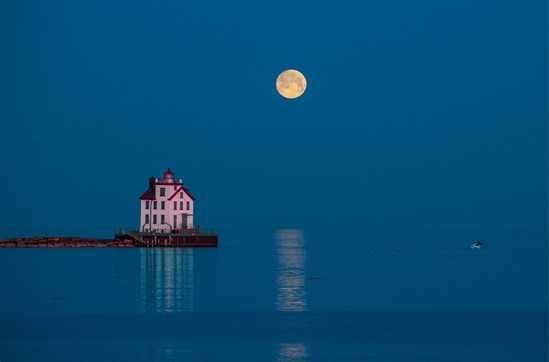 The full Moon hangs in the vibrant blue sky, above the Lorain Lighthouse. The sky and lake are the same shade of blue and blend into one another. The lighthouse is white with red roofs. It sits on a small piece of land that juts out into the water.