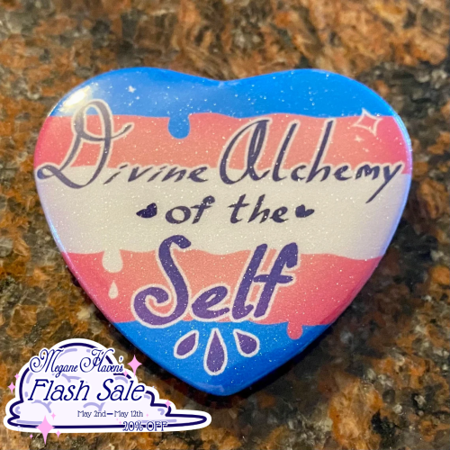 A subtly sparkly heart button with an illustrated trans pride flag background that says: "Divine Alchemy of the Self"