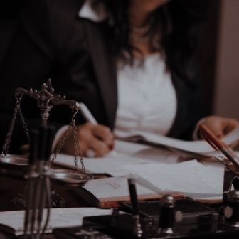 An image of a person signing a document with others littering the desk that has a scale of justice on it