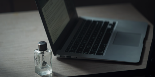 On a pale wood table is a silver laptop and a small, glass bottle of water adorned with a silver crucifix with a silver cap.