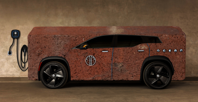 A manufacturer's publicity image of a Fisker Ocean electric SUV in a garage next to a wall-mounter charger. The car has been replaced by a gigantic, red clay brick.