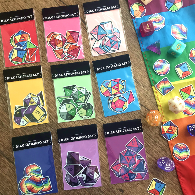 Promotional photo of the previously described dice sticker packs, arranged in rainbow order in a 3x3 grid. A rainbow pride flag is laid out beside the sticker packs with individual stickers and actual dice scattered across it.