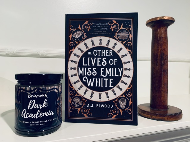 Shown is THE OTHER LIVES OF MISS EMILY WHITE (with cover art depicting a zoetrope image of a woman as well as corner images of a skull, flowers, an artist's palette, and a stack of books) on a shelf next to a Dark Academia candle and a loom spool. Photo by AHS.