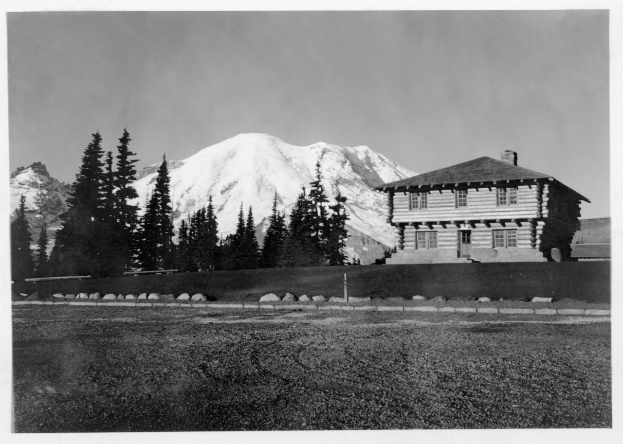 Black and white historic photo of a square two-story log building with a wood shingle roof next to a large dirt parking lot in front of a view of glacier-covered mountain.