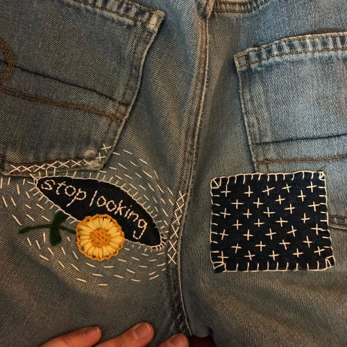 Embroidered patches covering up holes in a pair of jeans, including the words "Stop looking" and a yellow flower.