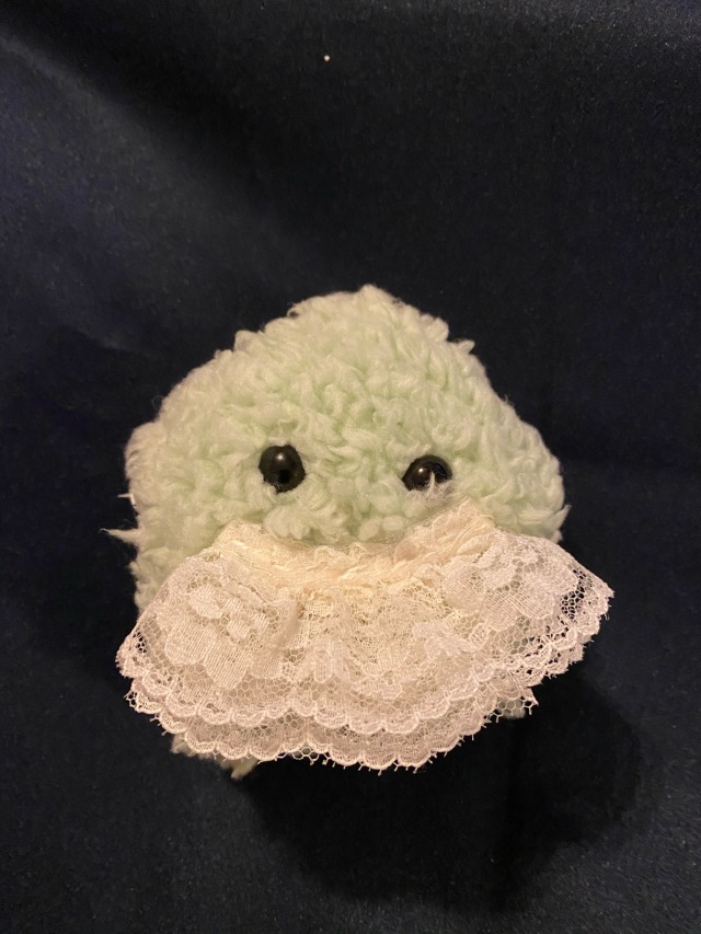 A small green star shaped plushie. it has shiny black eyes and a large scrap of lace making a mustache. it is made of puffy faux fur.