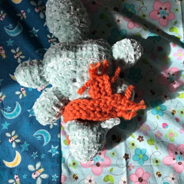 A blue knitted squirrel wearing an orange scarf sits on patterned blue fabric.