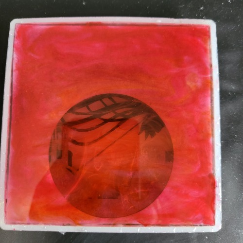 A layer of irregular deep red resin has been added to the topmost layer of the silicone mold. The layers of paper can be glimpsed through the circle cut-out in the bottom half of the square.