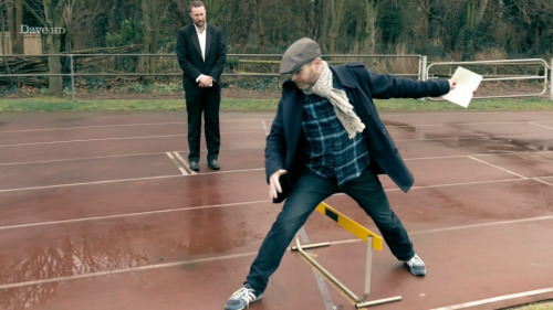 [ID: Five Screencaps Of Dave Gorman On Taskmaster, Traversing An Outdoor Athletics Track With Comically