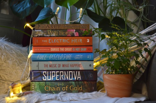 stack of books, on the left are fairylights, on the right is a plant and here are more plants in the background, the books are from top to bottom: aset, wild beauty, the electric heir, darius the great is not okay, full disclosure, the music of what happens, supernova, chain of gold