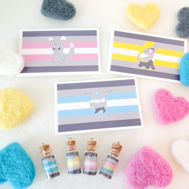 a photo of various demigender themed items including needle felted hearts, three stickers, a four bottle charms. The stickers feature the pokemon jangmo-o, carbink, and minccino.