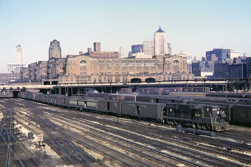 From Trains.com:“A Pennsylvania Railroad Geep pulls a string of express cars in front of railroad’s 