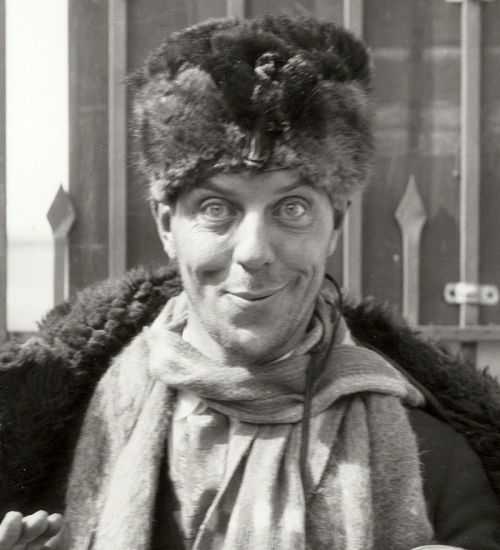 Karl Dane wearing and carrying a variety of clothes, including a fur hat, poses by the MGM studio ga