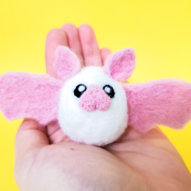 A photo of a white needle felted bat with a pink nose, ears, and wings. It is being held by a hand.